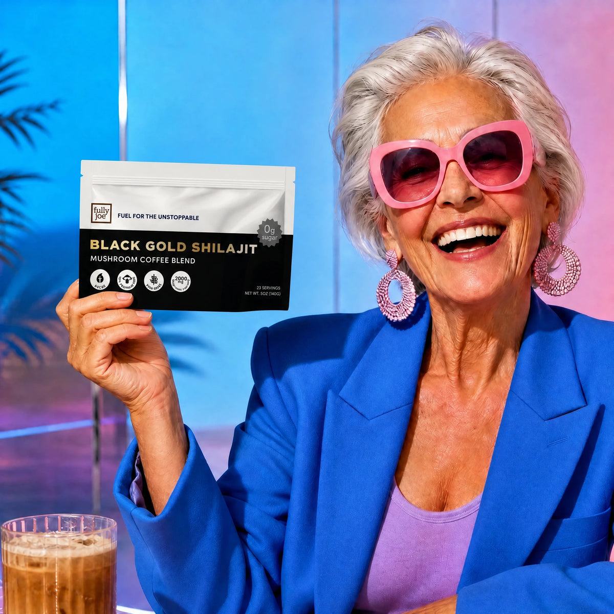 An older woman in pink sunglasses and a blue blazer smiles while holding a packet of Fully Nutrition Shilajit Coffee; an iced coffee from Fully Nutrition sits on the table.