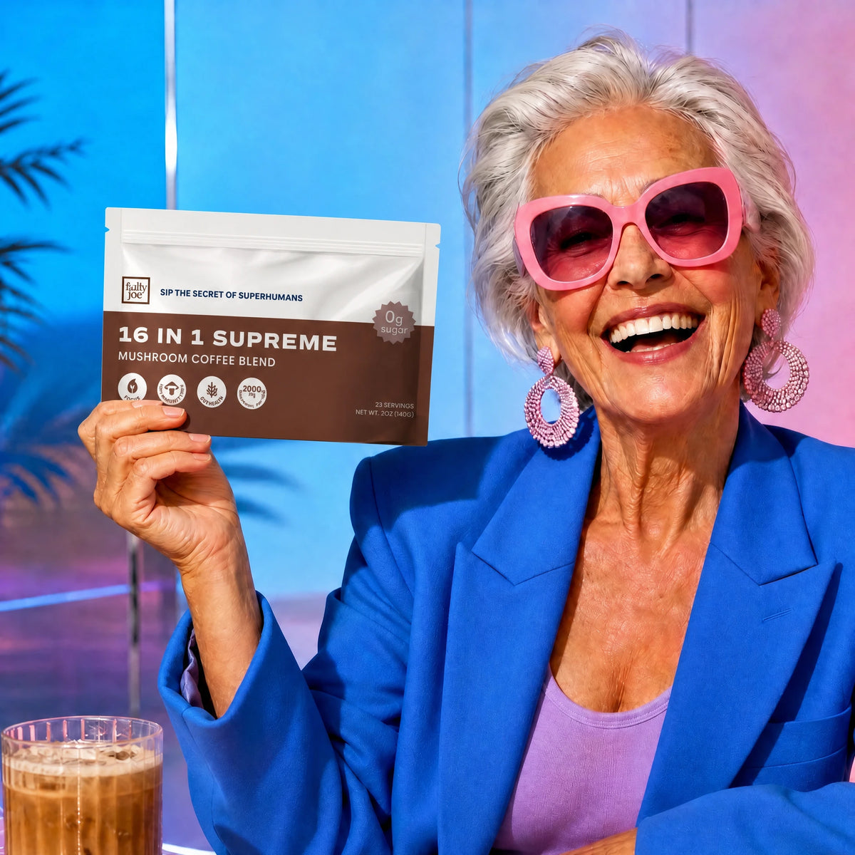 An older woman in pink sunglasses and a blue blazer smiles as she holds a packet of Fully Nutrition 16 in 1 Mushroom Blend Coffee near a glass of iced coffee, seated at a table with a vibrant background.