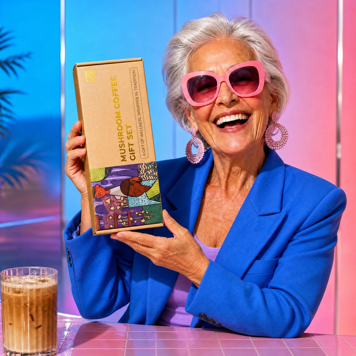 An older woman in a blue blazer and pink sunglasses smiles as she holds a box labeled "Mushroom Coffee Gift Set" from Fully Nutrition. An iced coffee rests on the table in front of her.