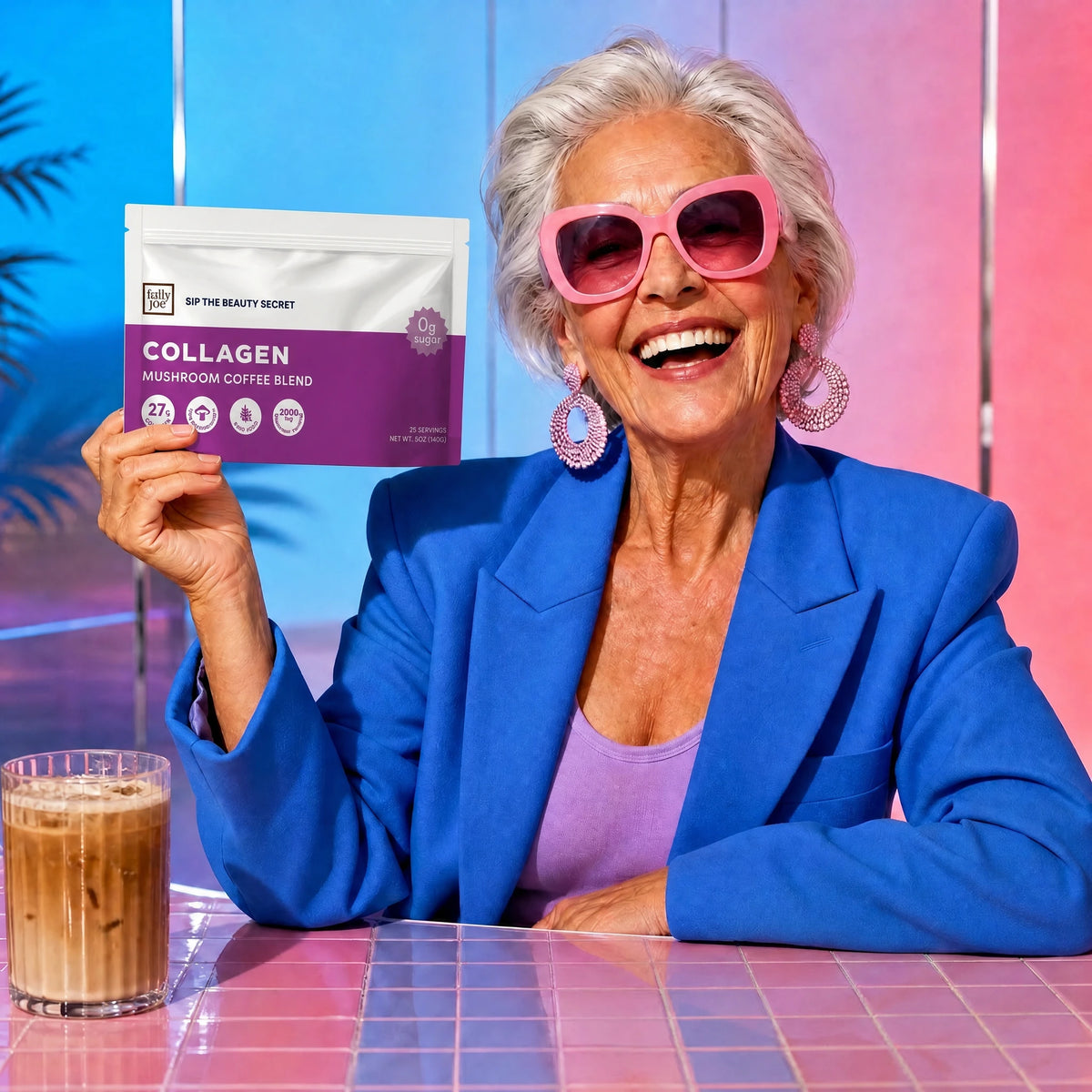 A stylish older woman in vibrant pink sunglasses and a blue blazer smiles as she holds a package of Fully Nutrition Collagen Mushroom Coffee by Fully Nutrition; an iced coffee rests on the table, symbolizing her embrace of age-defying beauty and brain-boosting benefits.