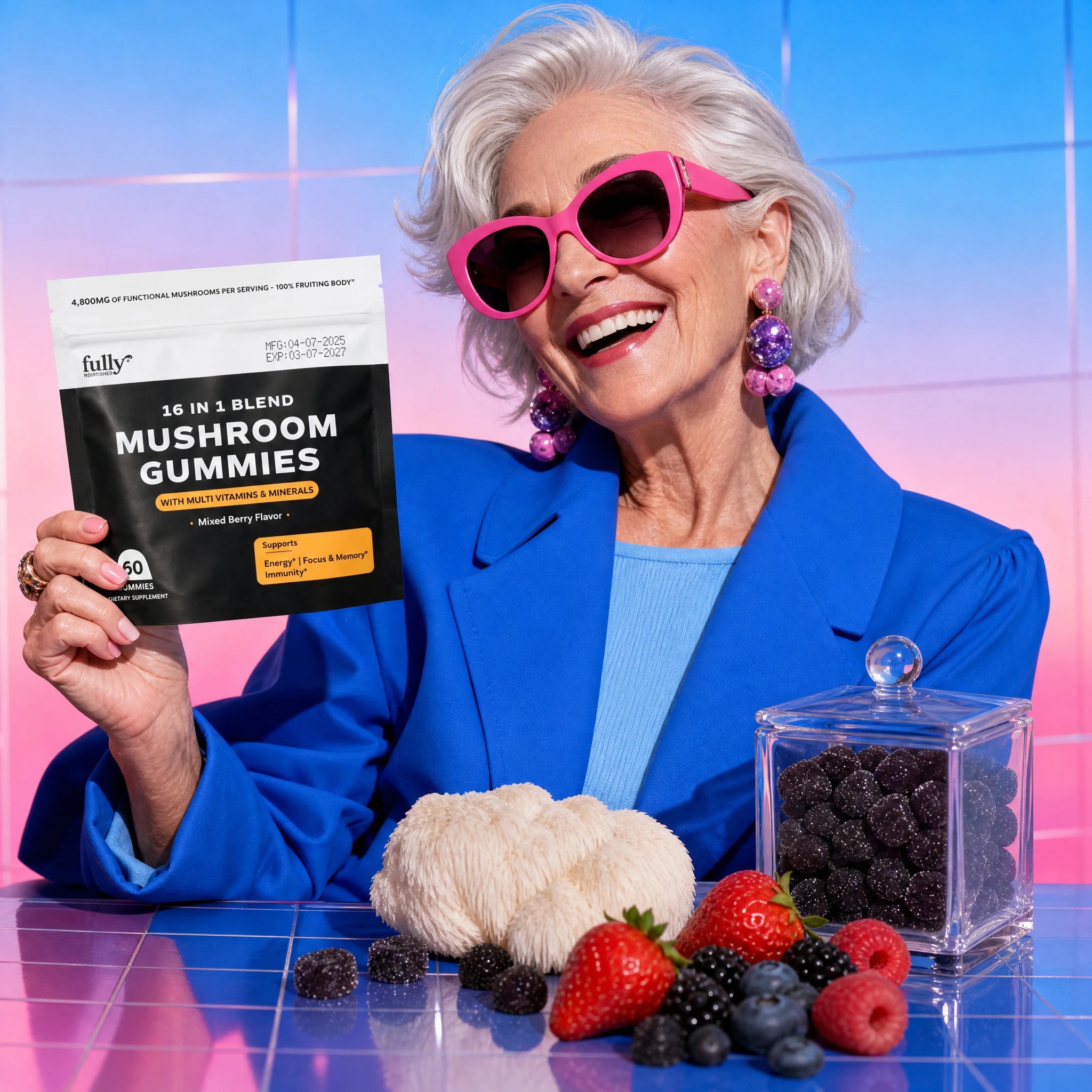 A smiling older woman in pink sunglasses holds a bag of Fully Nutrition 16 in 1 Mushroom Gummies by Fully Nutrition, with berries, mushrooms, and a glass container of gummies on the table against a colorful backdrop.
