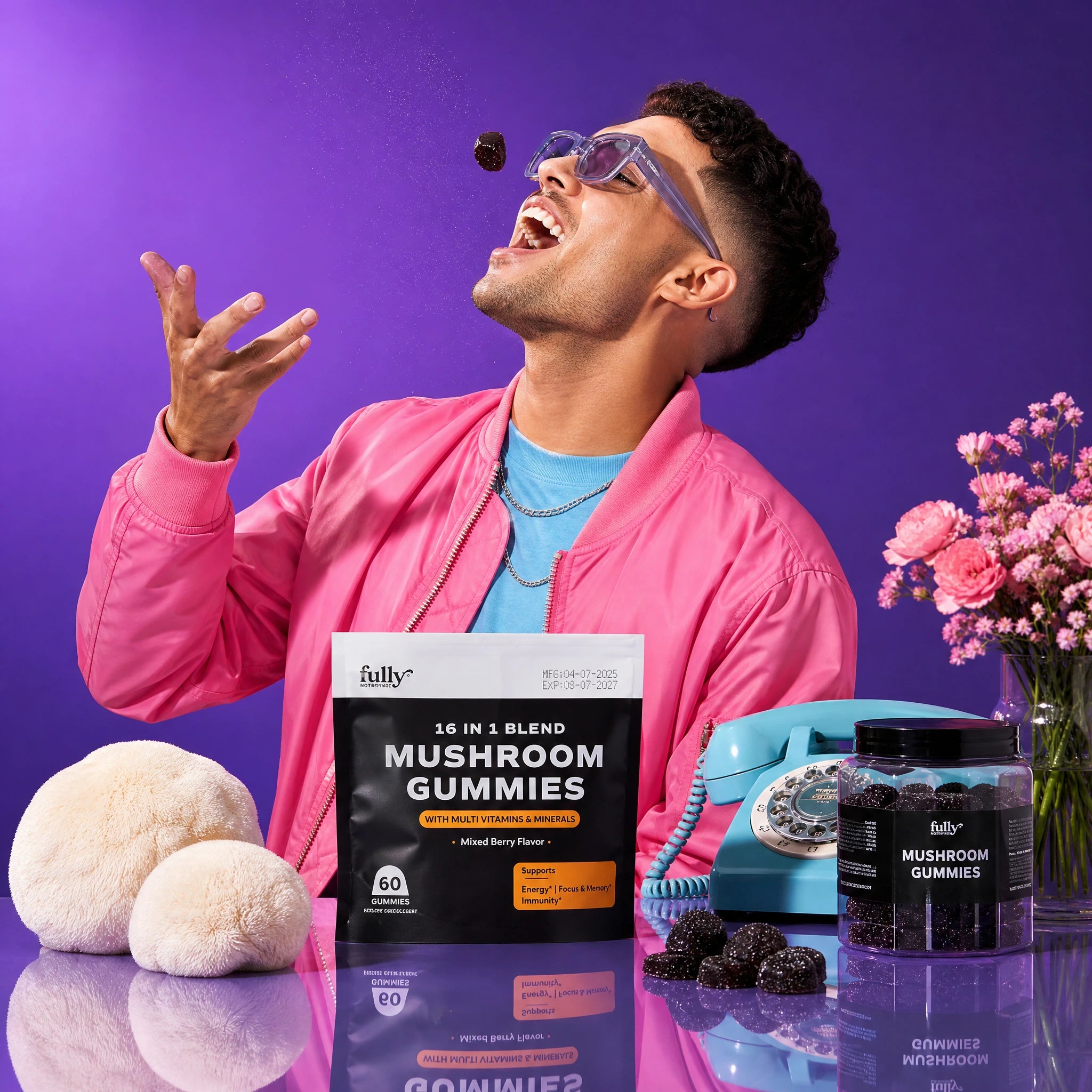 A man in a pink jacket tosses a gummy above his mouth, surrounded by Fully Nutrition 16 in 1 Mushroom Gummies packaging, a blue rotary phone, flowers, and mushroom props on a purple background.
